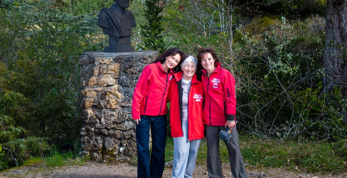 Trois femmes souriantes en vestes rouges posant près d'une statue de buste à Meyrueis en Lozère 48